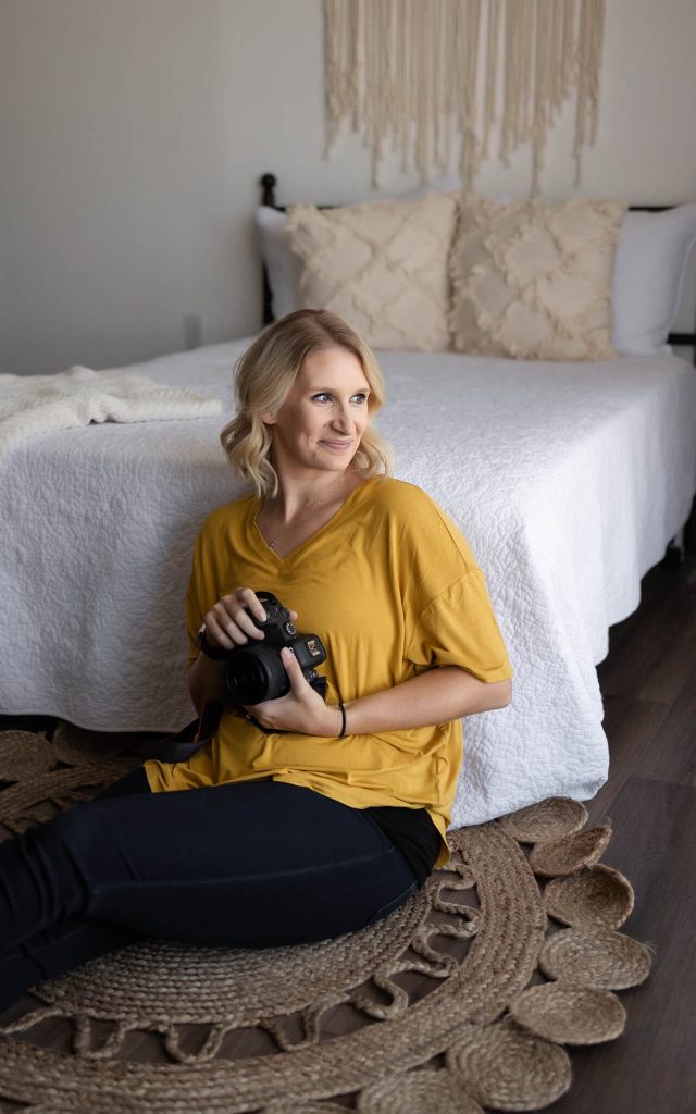 Brittany Moncrieff photographer in yellow shirt holding camera on woven rug, cozy bedroom with white bed and decorative pillows | Maryville maternity photographer