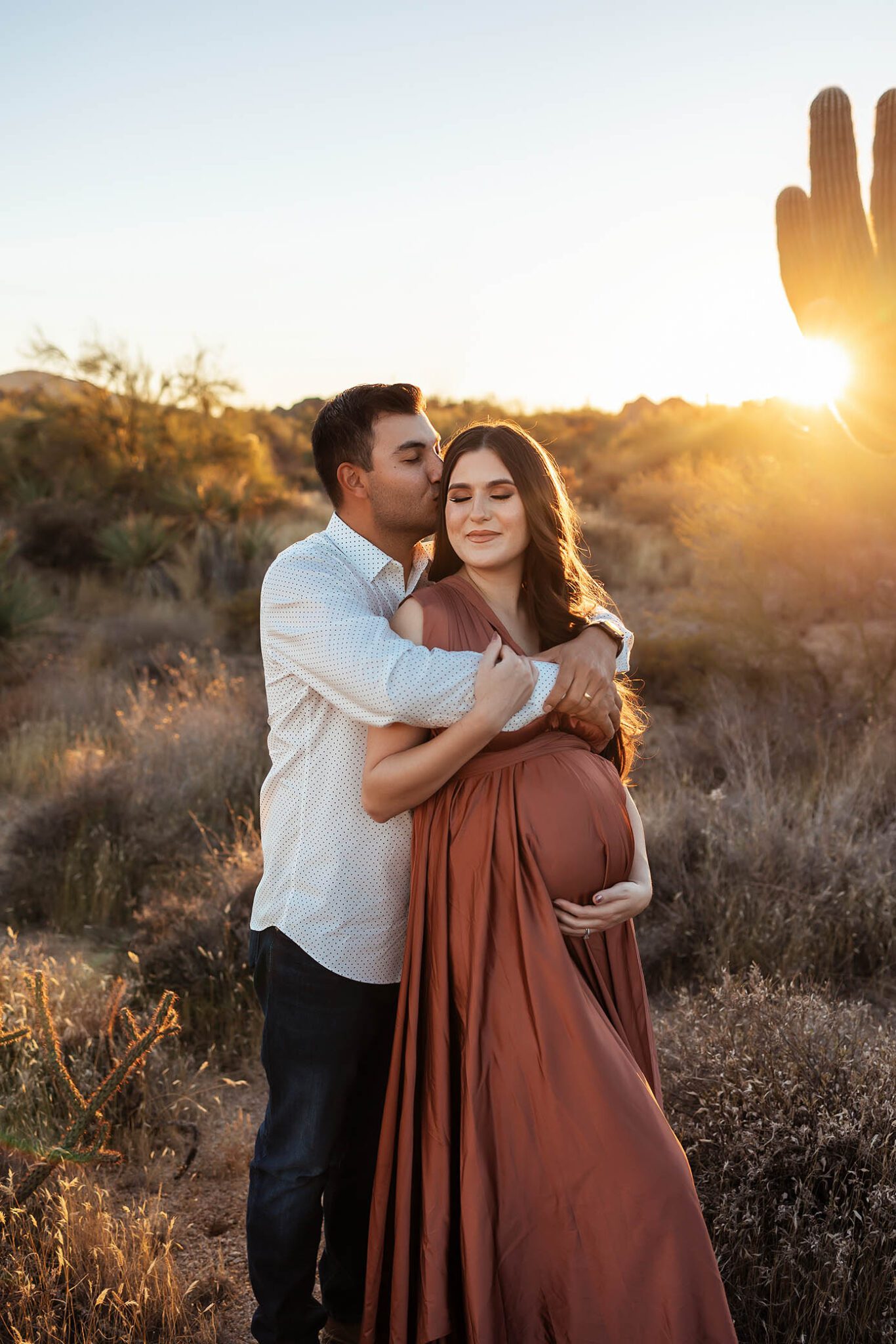 Outdoor Maternity Photography in Tennessee - Man embraces and kisses pregnant woman's forehead at sunset in desert, large cactus in background.