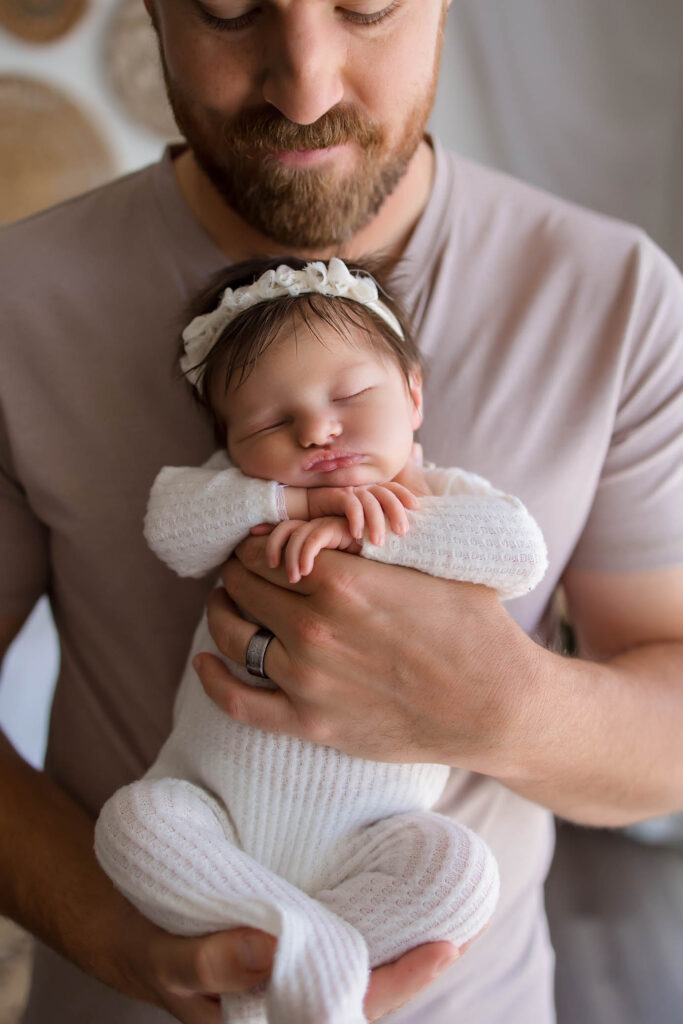 Bearded man cradles sleeping newborn in white and floral headband against his chest.