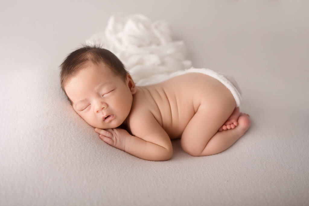 Newborn baby in a diaper sleeps on a soft, light surface with a white blanket background.