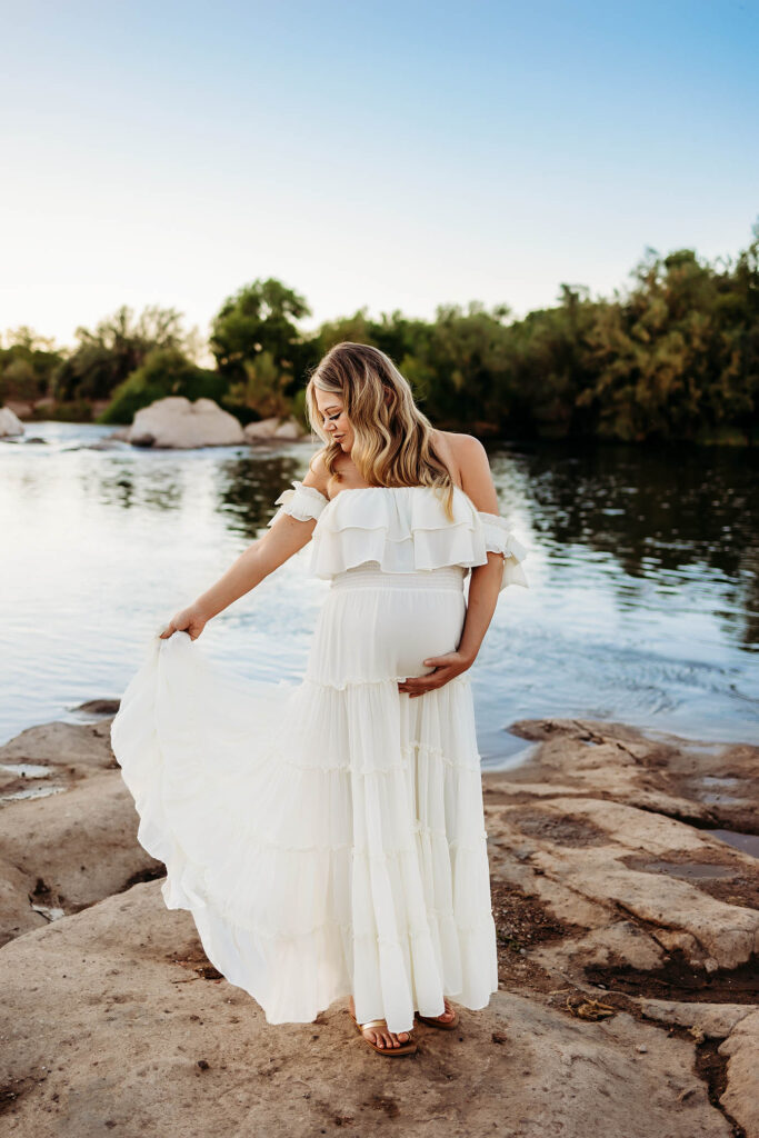 Pregnant woman in white dress stands on riverside rocks, holding her belly, trees and blue sky behind.