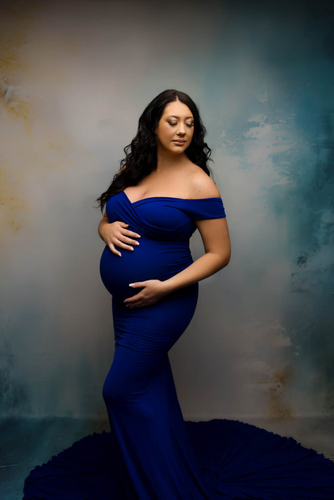 Pregnant woman in royal blue off-shoulder gown cradling belly, standing before blue-beige backdrop.