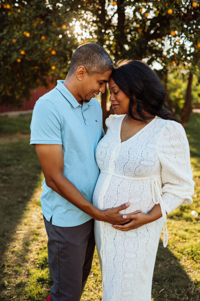 Smiling couple outdoors, foreheads touching; pregnant woman in white dress holds belly, man in blue shirt.
