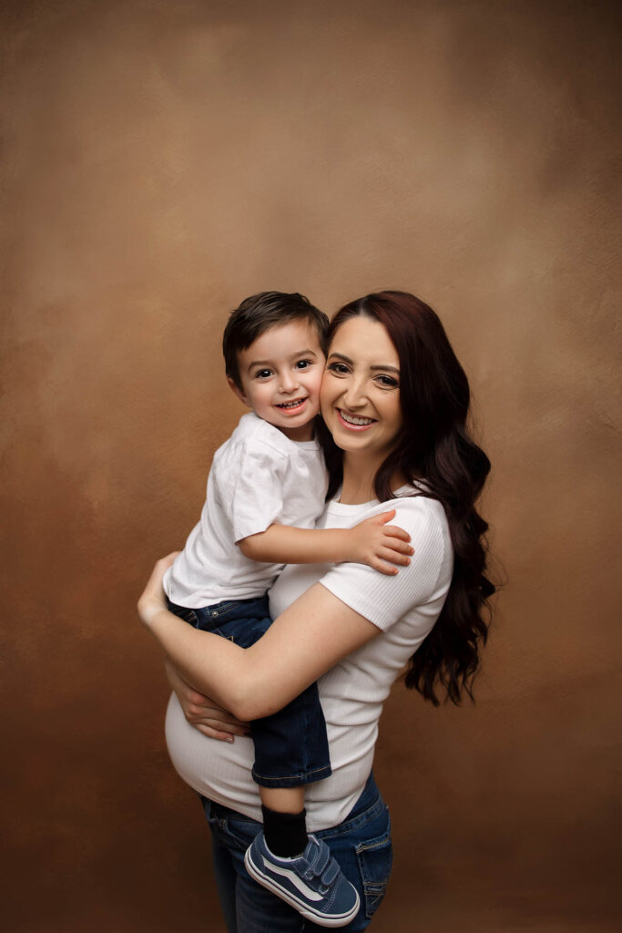 Woman with long brown hair holding smiling boy, both in white shirts and jeans, brown studio backdrop.