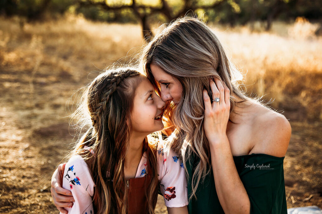 Woman and young girl smiling, touching foreheads outdoors amid dry grass and trees.