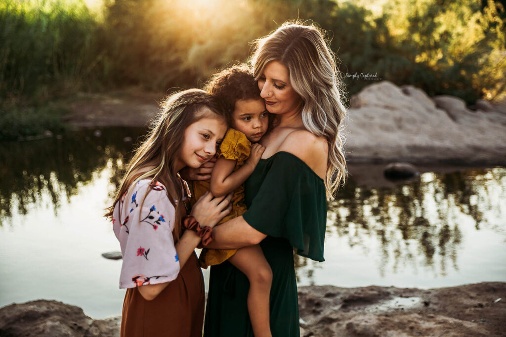 Woman holding a young child and older girl by a riverbank at sunset, standing close together.