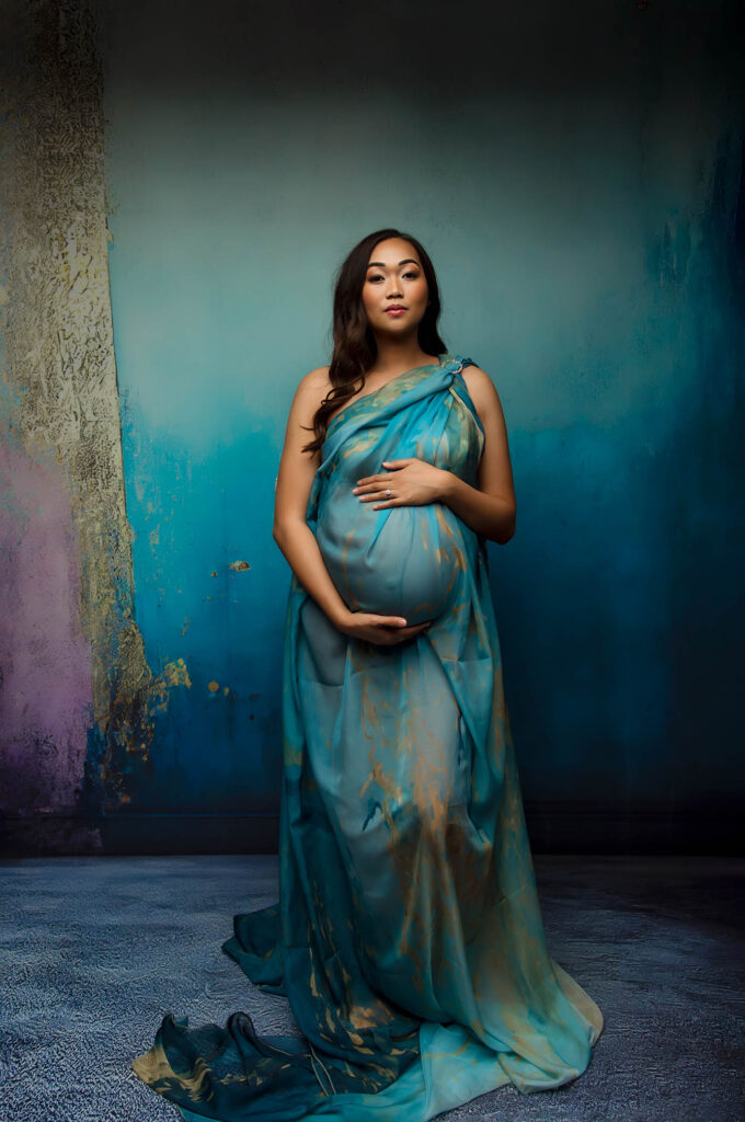 Pregnant woman in blue and gold gown, hands on belly, stands before textured blue and gold backdrop.