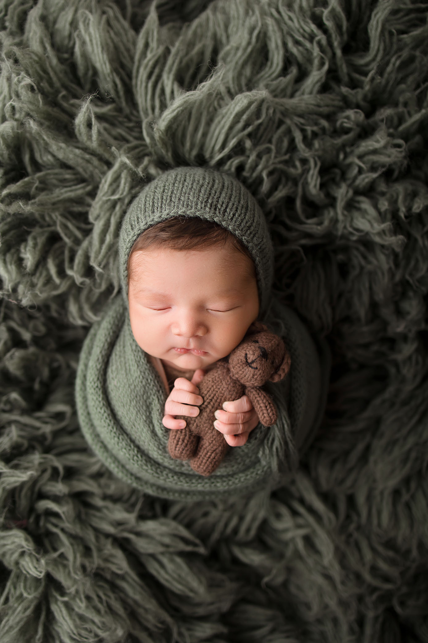 Newborn in green knit blanket and hat, lying on textured green fabric, holding small brown bear.