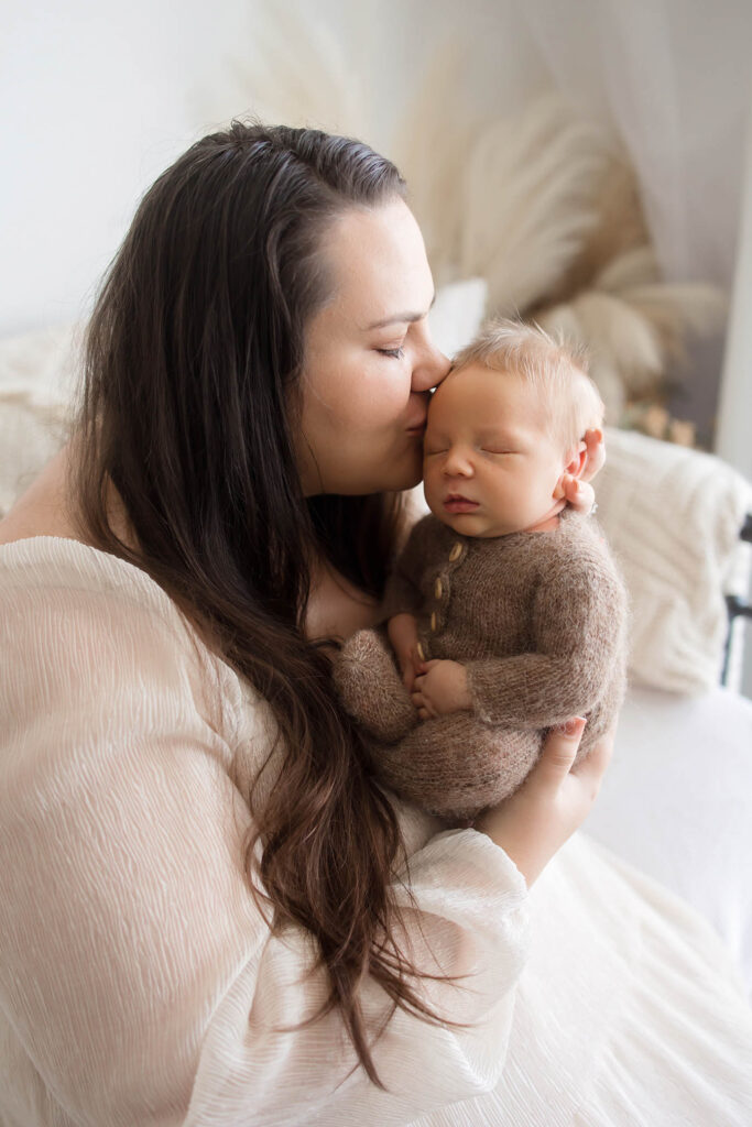 Woman with long brown hair kisses sleeping newborn in brown knit outfit, holding baby indoors.