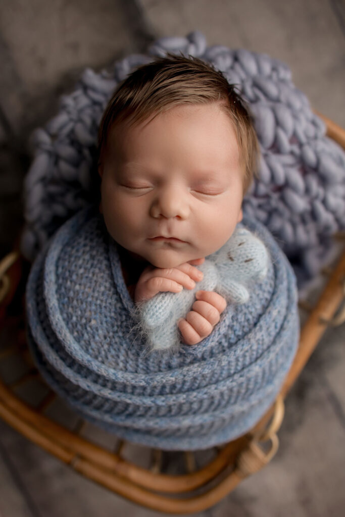 Newborn baby swaddled in blue knit blanket, eyes closed, cuddling small blue stuffed toy on cushion.