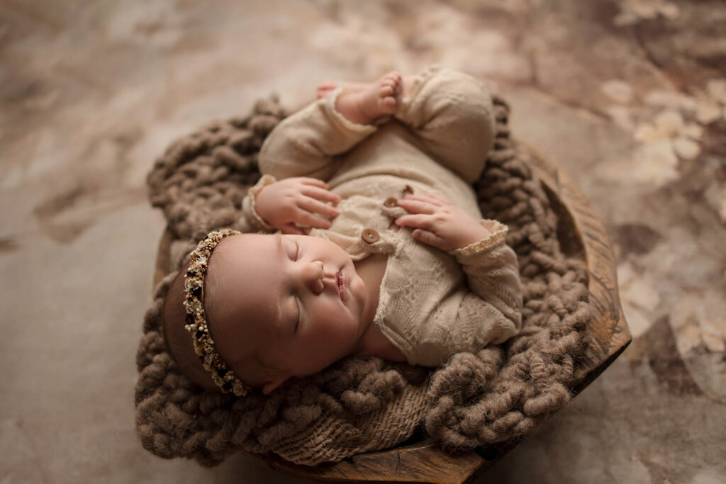 Newborn baby in beige outfit and floral headband sleeps on a chunky knit blanket in wooden bowl.