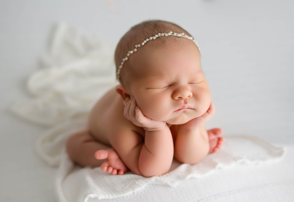 Sleeping newborn with closed eyes, hands under chin, on white blanket, wearing a delicate headband.