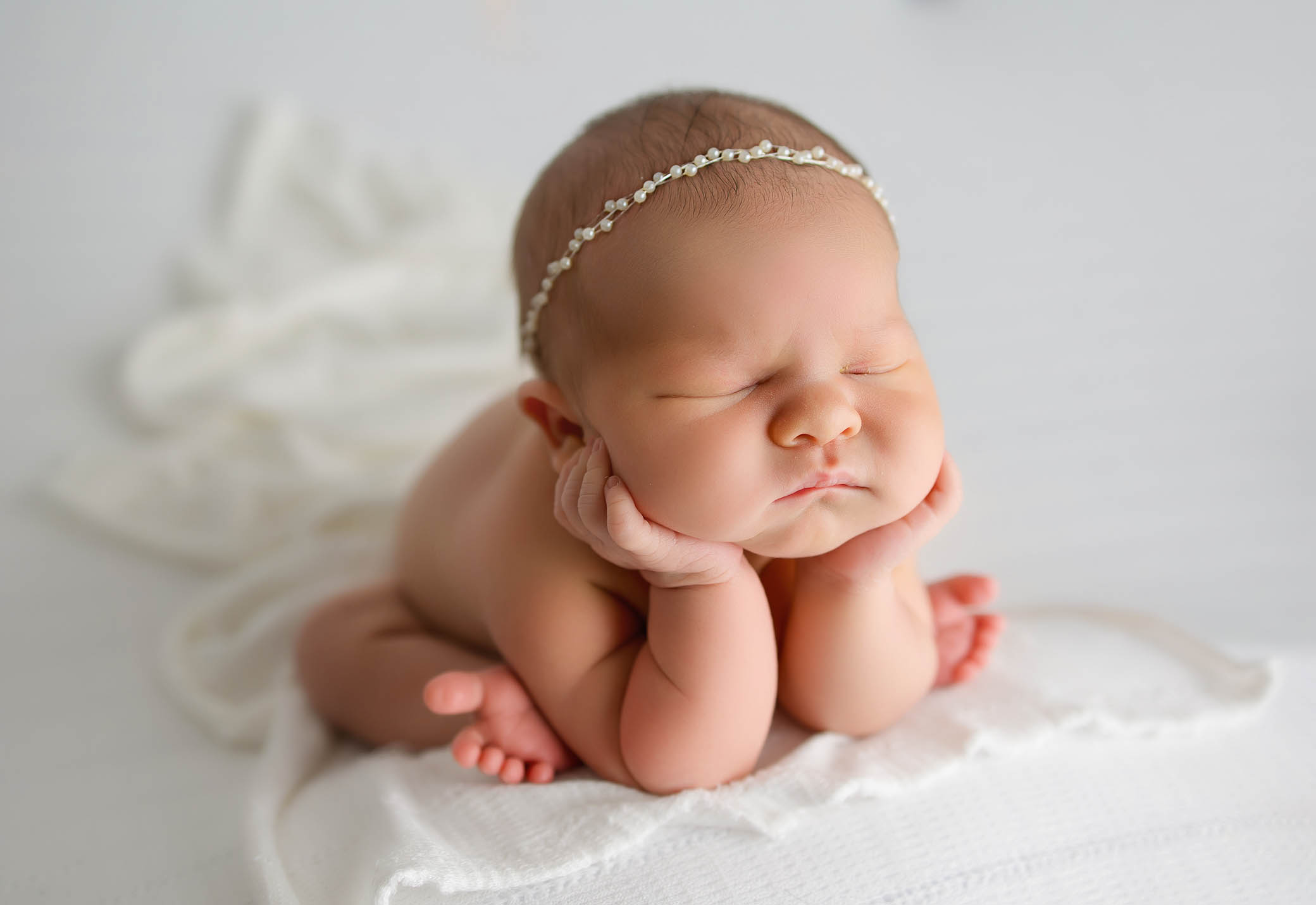Sleeping newborn with closed eyes, hands under chin, on white blanket, wearing a delicate headband.