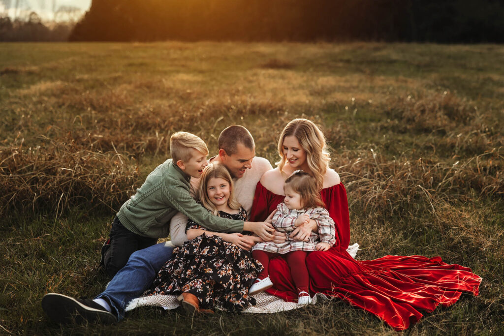 Moncrieff family of five enjoys sunset on a blanket in a grassy field, smiling and interacting together.