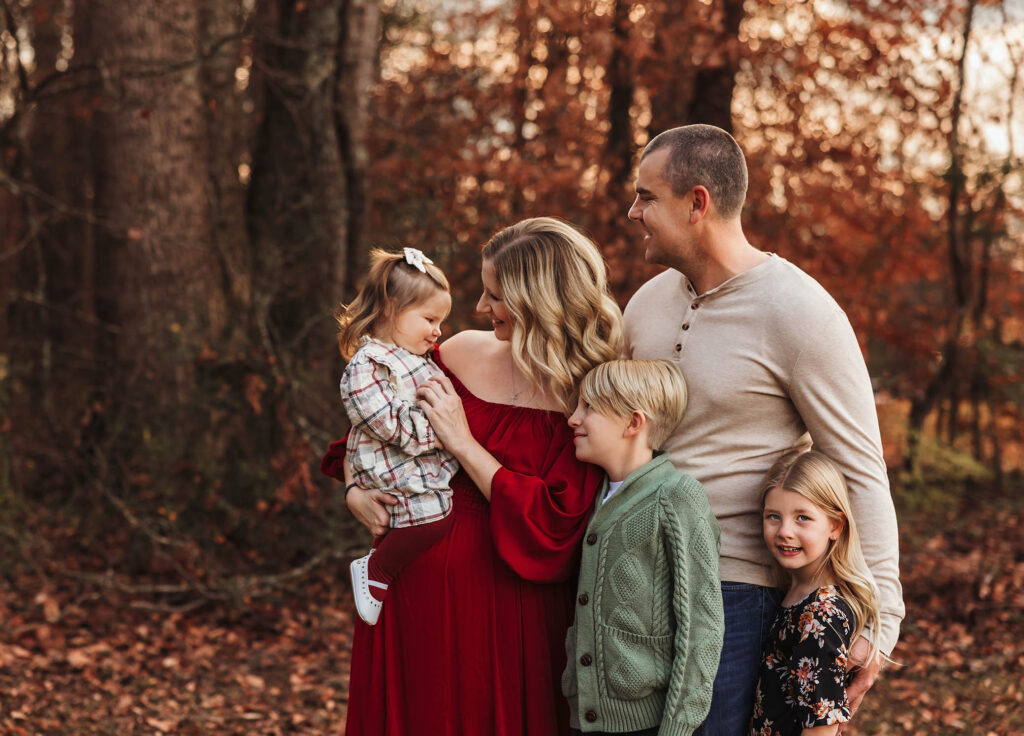 Smiling Moncrieff family of five stands together outdoors in autumn among fallen leaves and colorful trees.
