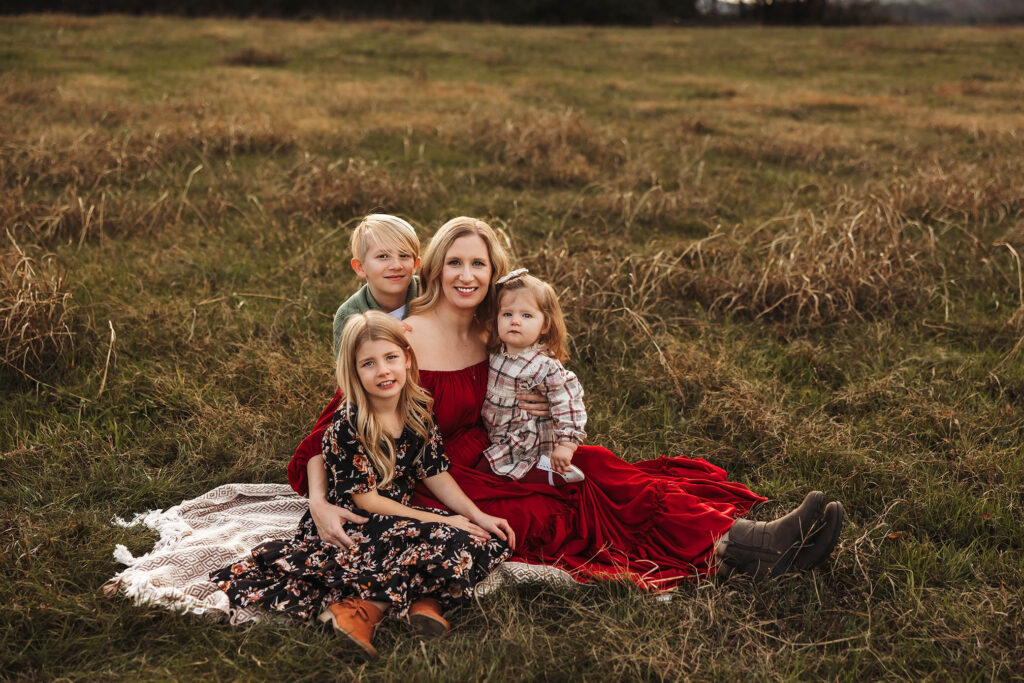 Brittany Moncrieff in red dress on picnic blanket in field with her three smiling children, two girls and a boy.