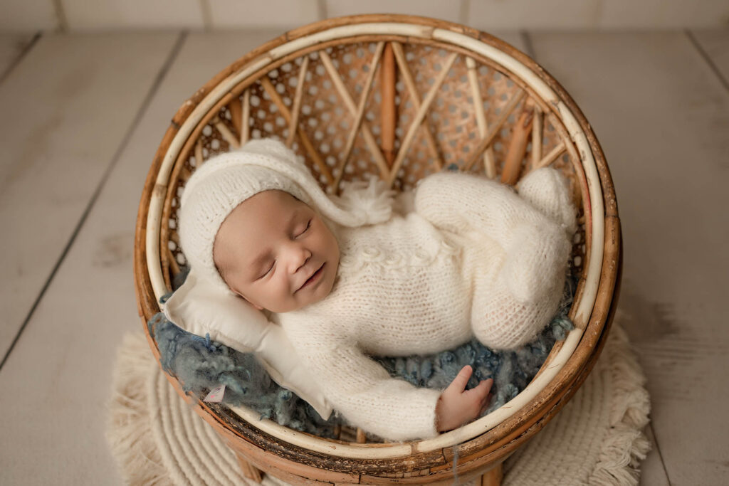 Smiling newborn in white knit outfit and hat, sleeping in wicker basket with pillow and blanket.