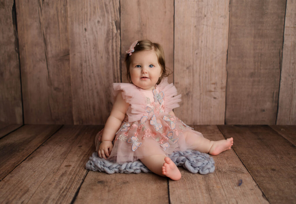 Baby in a pink butterfly dress sits on a gray blanket with a wooden background.