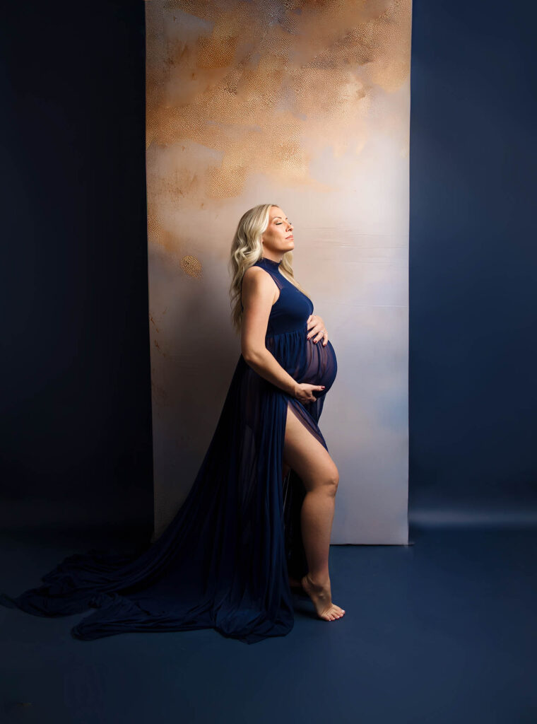 Pregnant woman in dark blue dress stands barefoot, holding belly and gazing upward against textured backdrop.