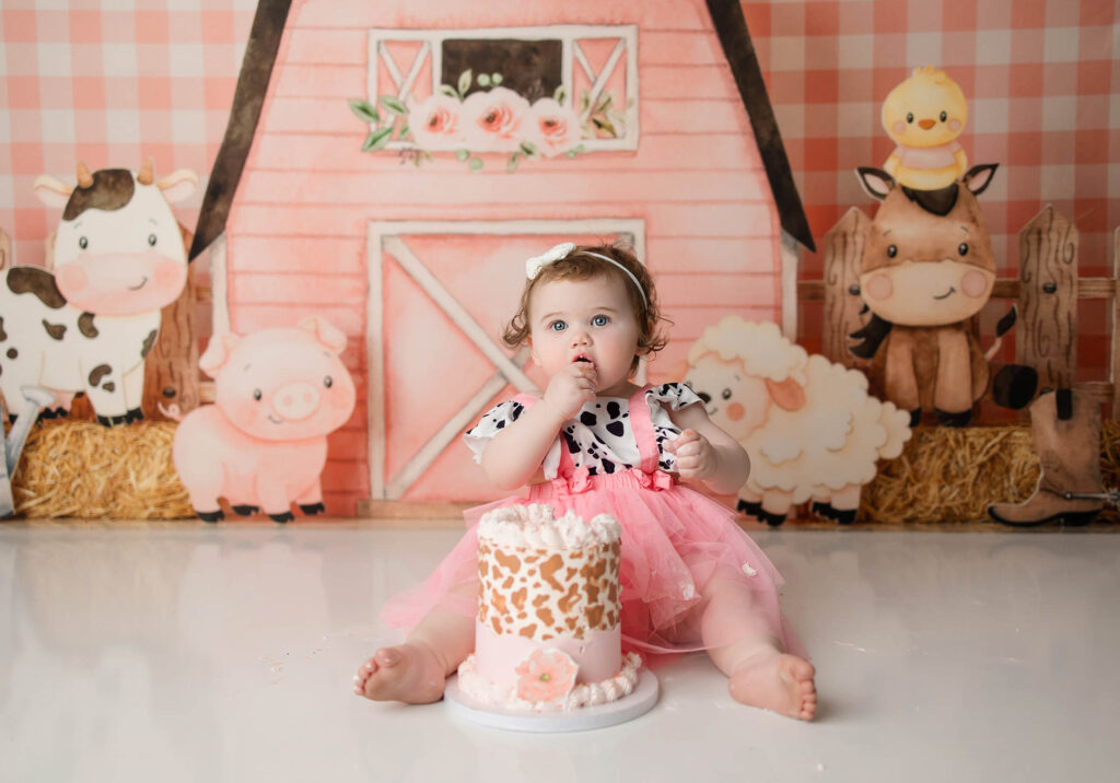 Baby in pink cow-print outfit eats cake on floor, with farm-themed backdrop of cartoon animals and barn.