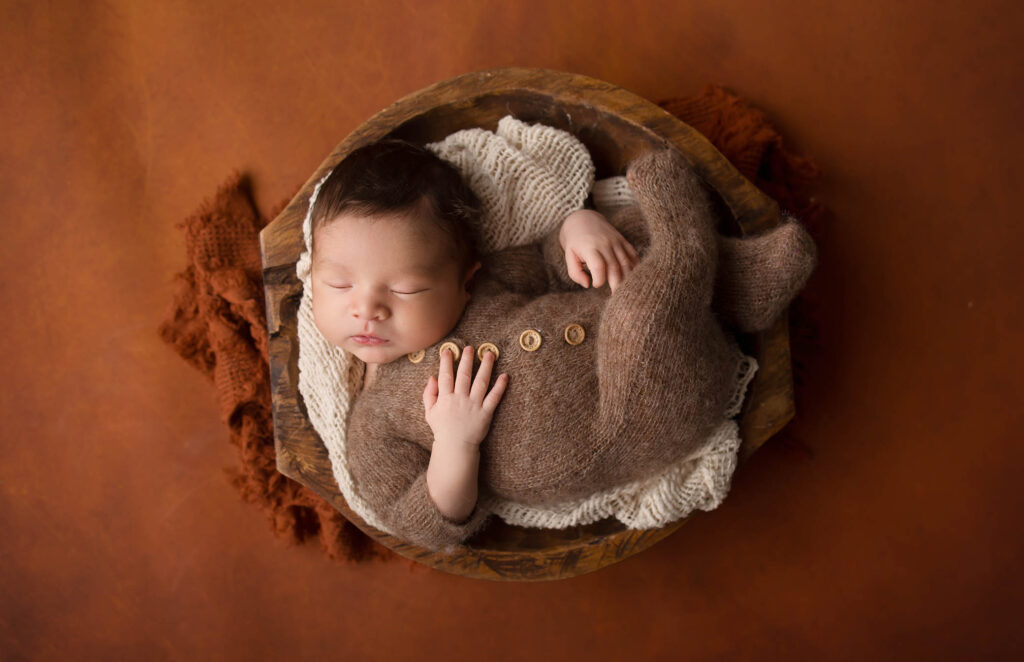 Newborn in brown knit onesie sleeps curled in wooden bowl, cozy blankets, brown background.