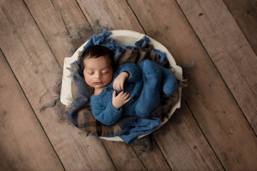Sleeping baby in blue outfit nestled in a white bowl on brown and blue blanket, wooden floor background.
