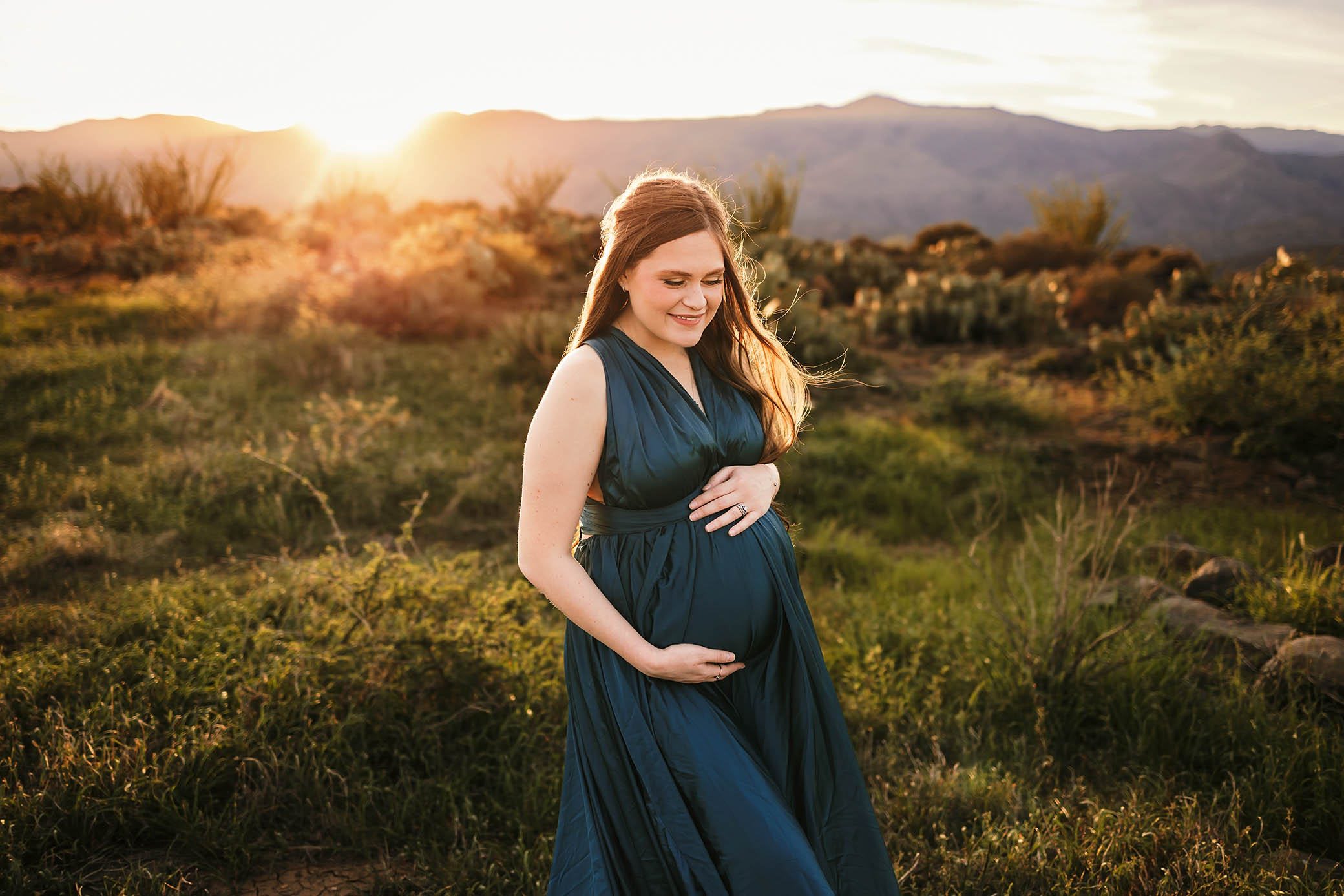 Pregnant woman in dark green dress smiling, holding belly in grassy field at sunset with mountains | Maryville maternity photographer