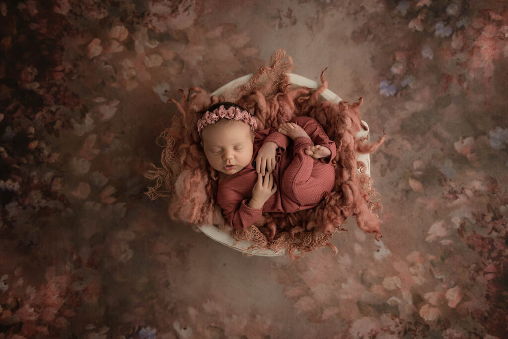 Sleeping newborn in mauve blanket and headband, nestled in a white bowl on floral-textured surface.