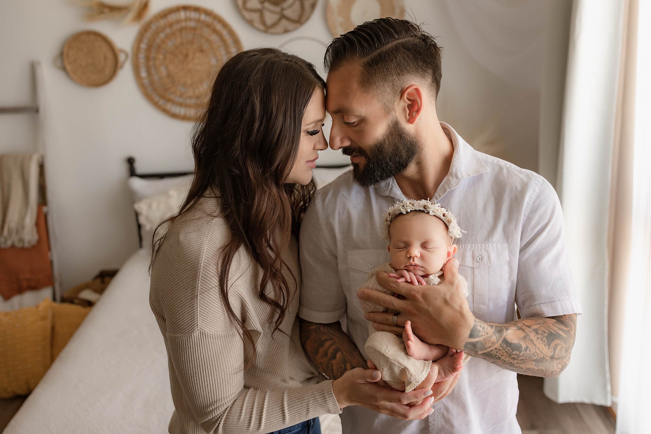 Couple holds sleeping baby in floral headband, with woven wall decor and soft indoor lighting.