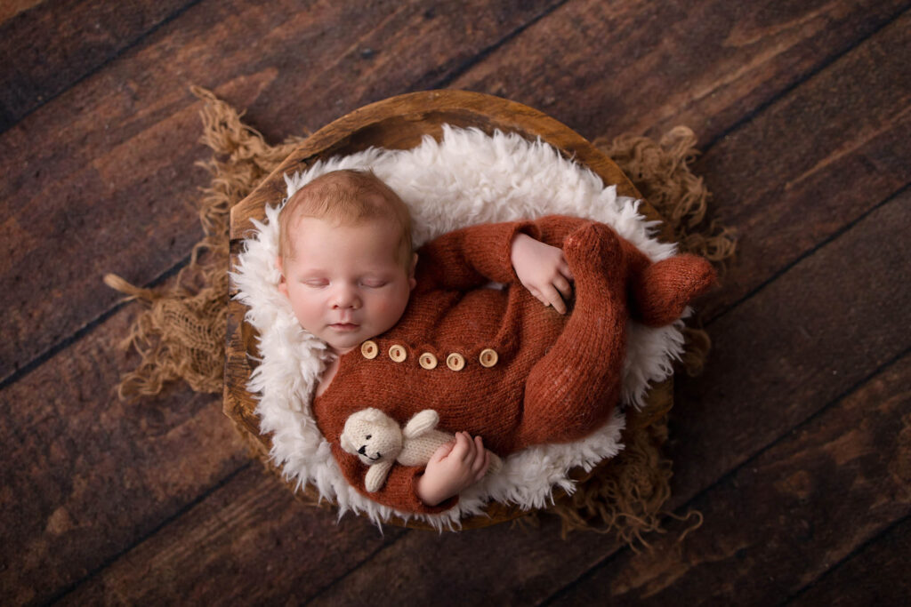Newborn baby in brown knit sleeps in a fur-lined basket, holding a stuffed bear on wooden floor.