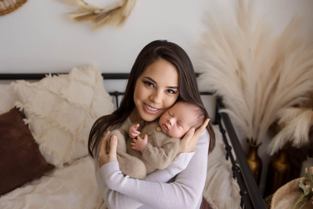 Smiling woman on bed holds sleeping baby in beige outfit, decorative pillows and pampas grass behind.