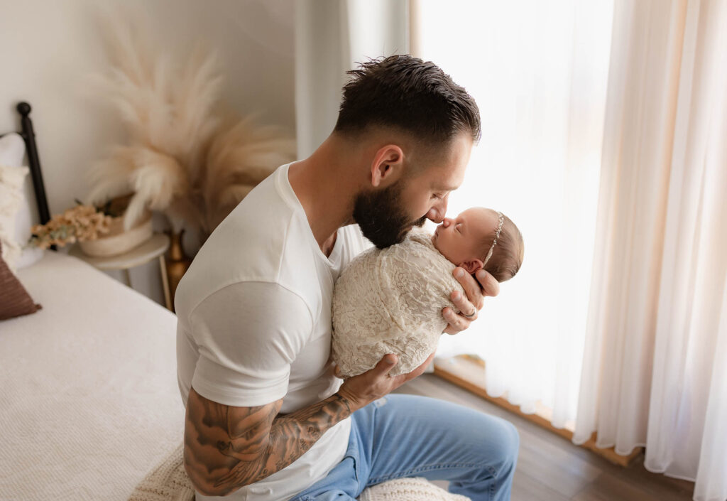 Tattooed man cradles swaddled newborn, touching noses on a bed in a softly lit room.