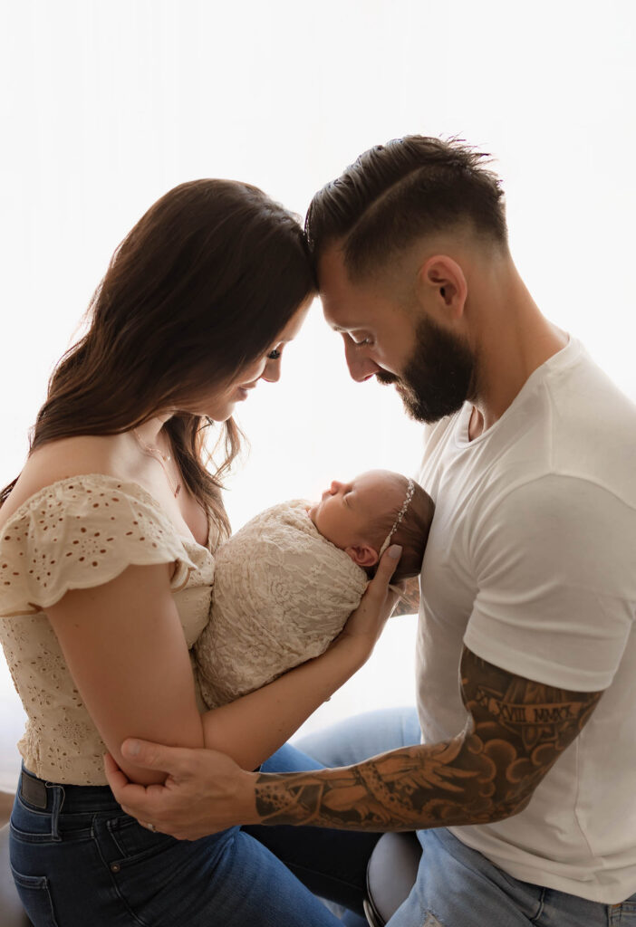 A couple sits forehead to forehead, lovingly holding their swaddled newborn baby between them.