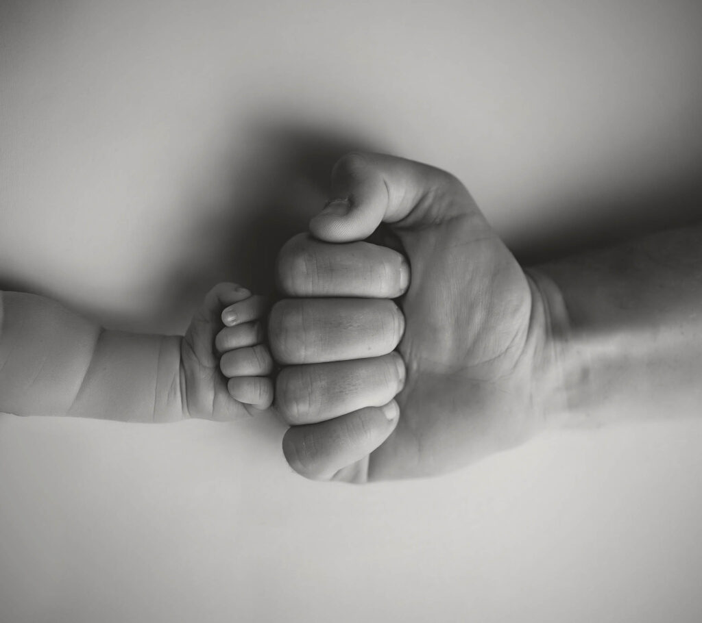 Baby’s hand holding adult finger, both resting on a flat surface in black and white.