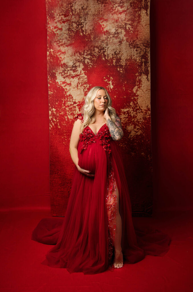 Pregnant woman in red semi-sheer gown poses against textured red and gold backdrop, hand on belly.
