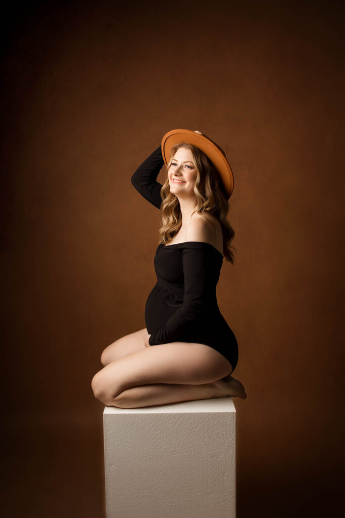 Smiling pregnant woman in black off-shoulder bodysuit and orange hat sits on white cube, brown backdrop.