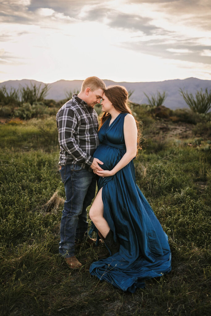 Man and pregnant woman in blue dress touching foreheads in grassy field at sunset, holding her belly.