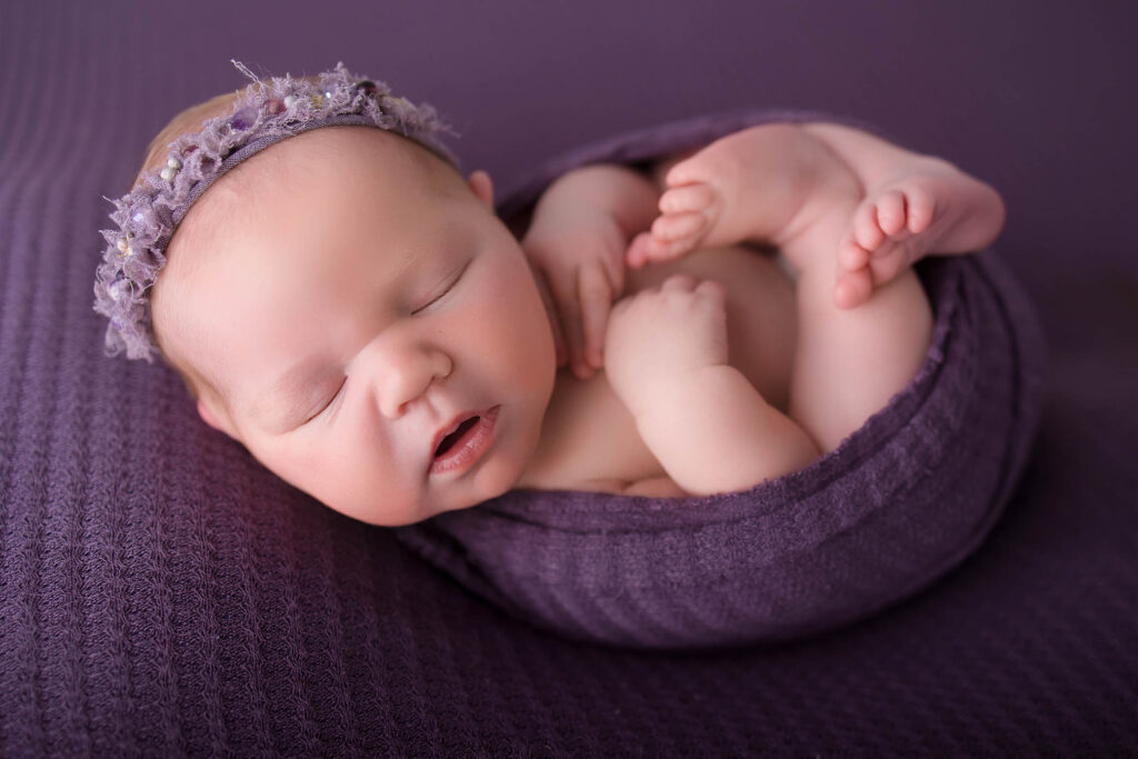 Sleeping newborn wrapped in purple cloth on textured blanket, wearing a delicate purple headband.