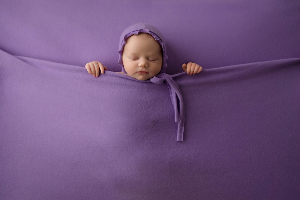 Newborn baby sleeping on a soft purple blanket, wearing a matching bonnet, hands near face.