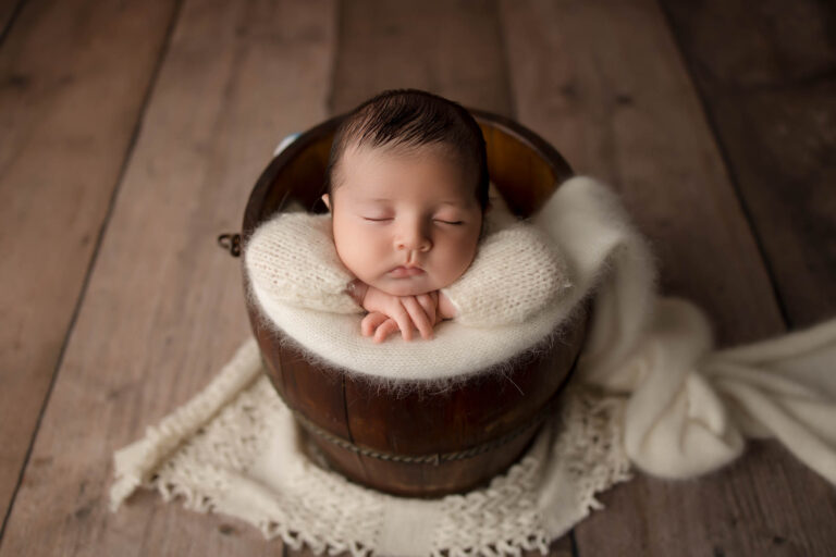 Newborn baby sleeps in a wooden bucket, wrapped in white blanket on lace mat and wooden floor.