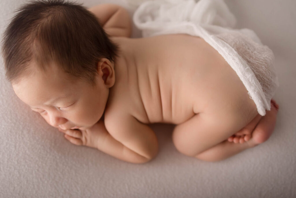 Newborn baby on stomach, resting on soft white surface, partially draped with sheer white cloth.