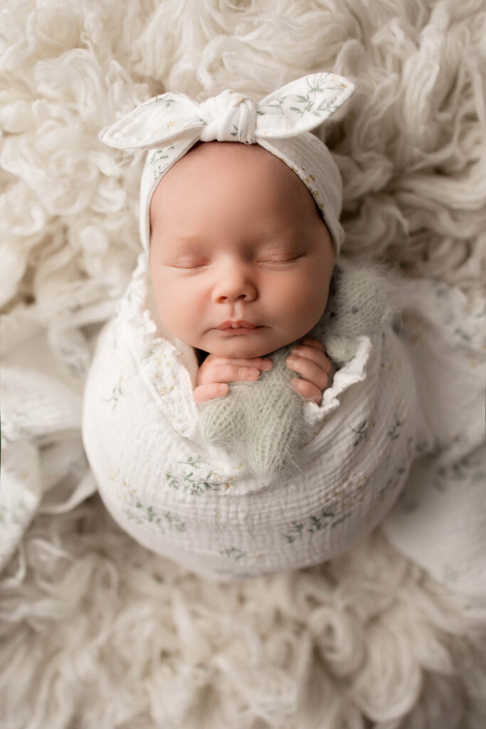 Newborn baby sleeping peacefully, wrapped in a white blanket with matching headband on fluffy surface.
