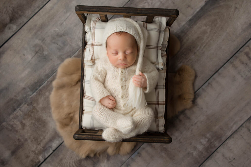 Newborn in white knitted outfit sleeps on wooden bed with plaid cushion, set on wooden floor.