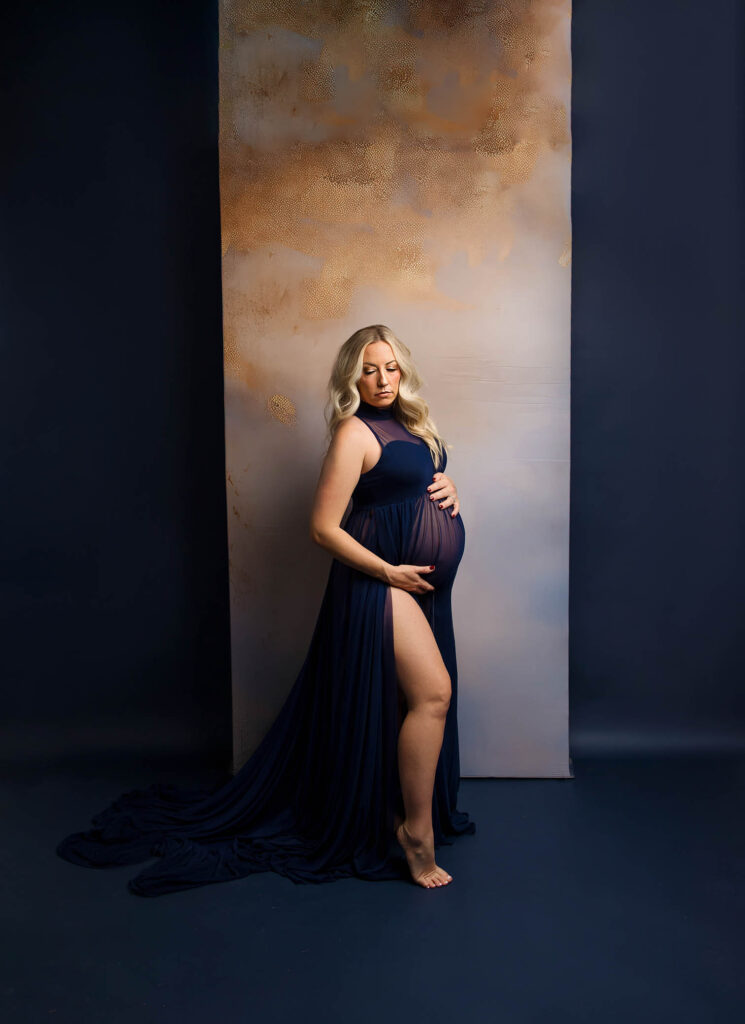 Pregnant woman in dark blue dress, barefoot, holding belly and looking down against soft lighting.