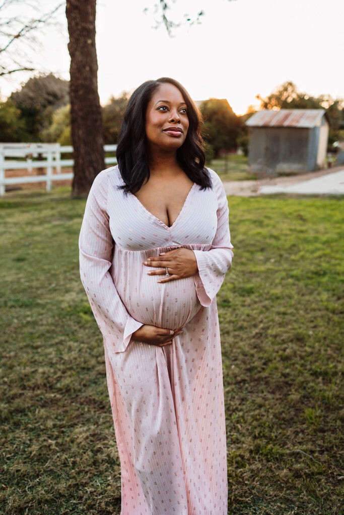 Pregnant woman in a long pink dress stands on grass, cradling her belly and gazing upward.