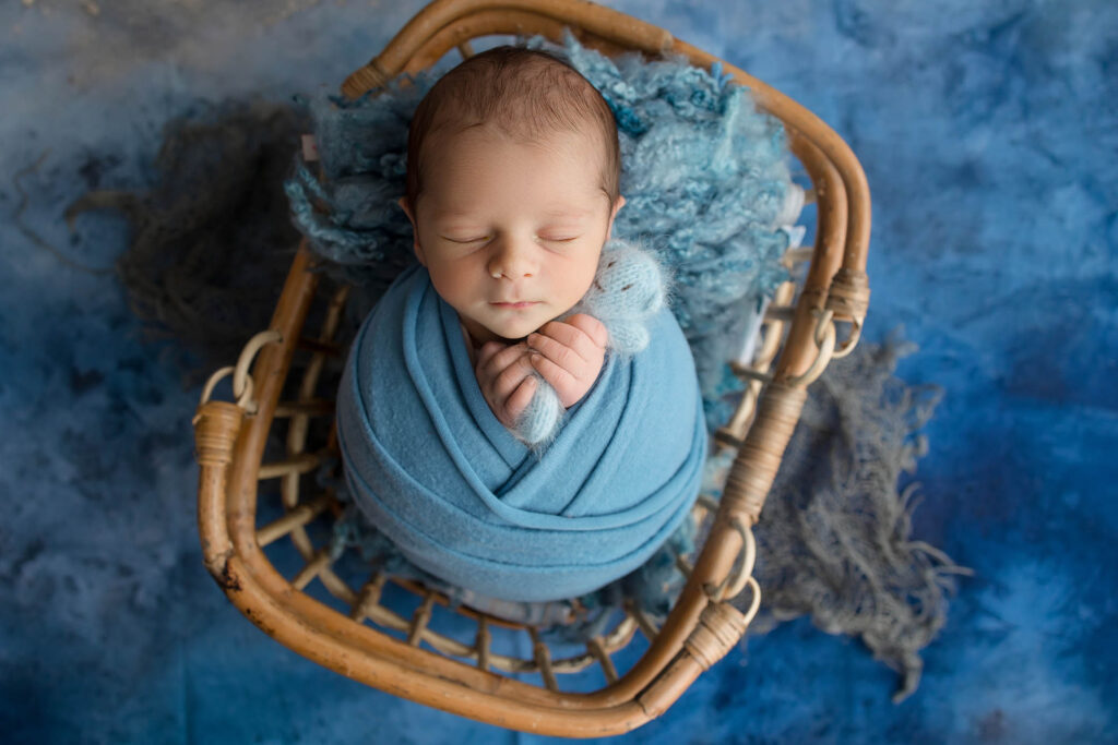 Sleeping newborn swaddled in blue blanket, nestled in wicker basket on blue textured backdrop.