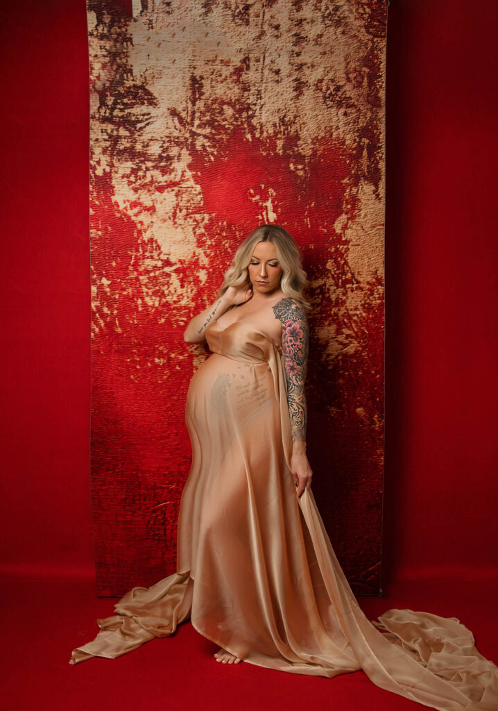 Pregnant woman in sheer gown stands barefoot, looking down against a textured red and gold background.