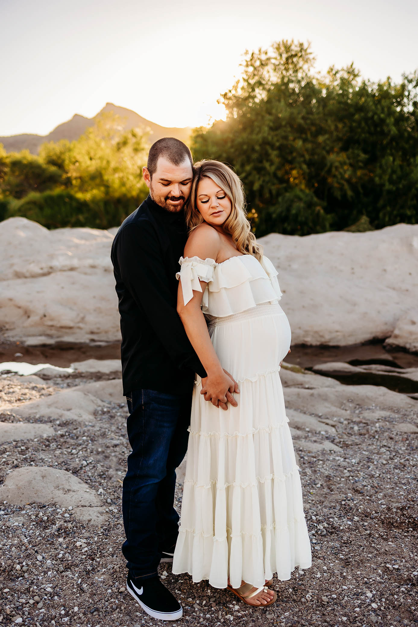 Pregnant woman in white dress embraced by partner outdoors, both looking down lovingly.