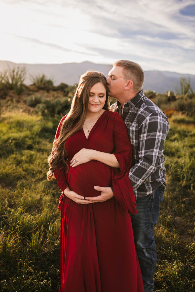 Man embraces pregnant woman in red dress, kissing her forehead outdoors with grassy field and mountains.