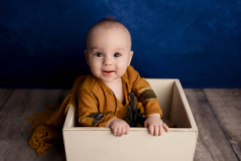Smiling baby in mustard yellow outfit sits in white box on wooden floor with blue background.